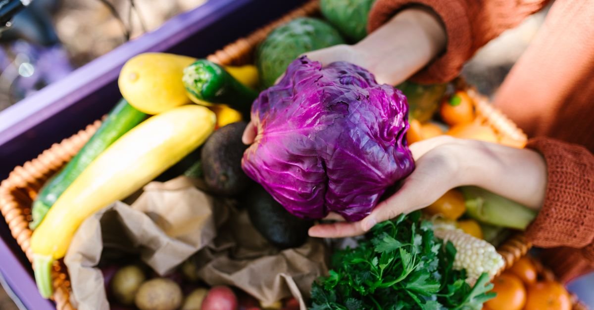 Mãos seguram repolho roxo sobre cesta de vime cheia de hortaliças e legumes frescos