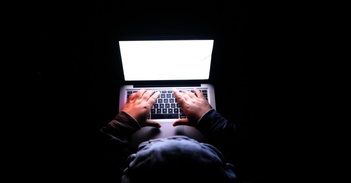 Mãos digitam em um notebook em ambiente escuro, com a tela iluminada