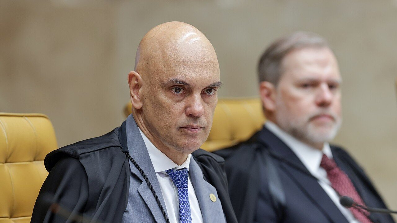 O ministro Alexandre de Moraes, de toga, sentado em sessão do plenário do Supremo Tribunal Federal, com o ministro Dias Toffoli desfocado ao fundo