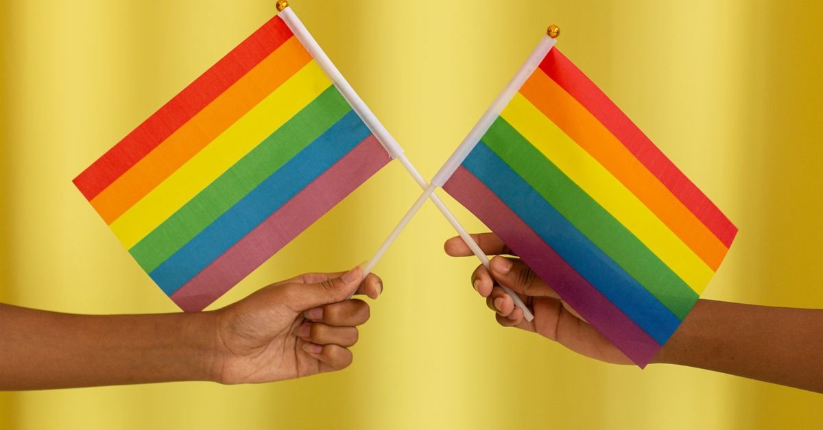 Duas mãos seguram bandeiras do arco-íris cruzadas diante de fundo amarelo