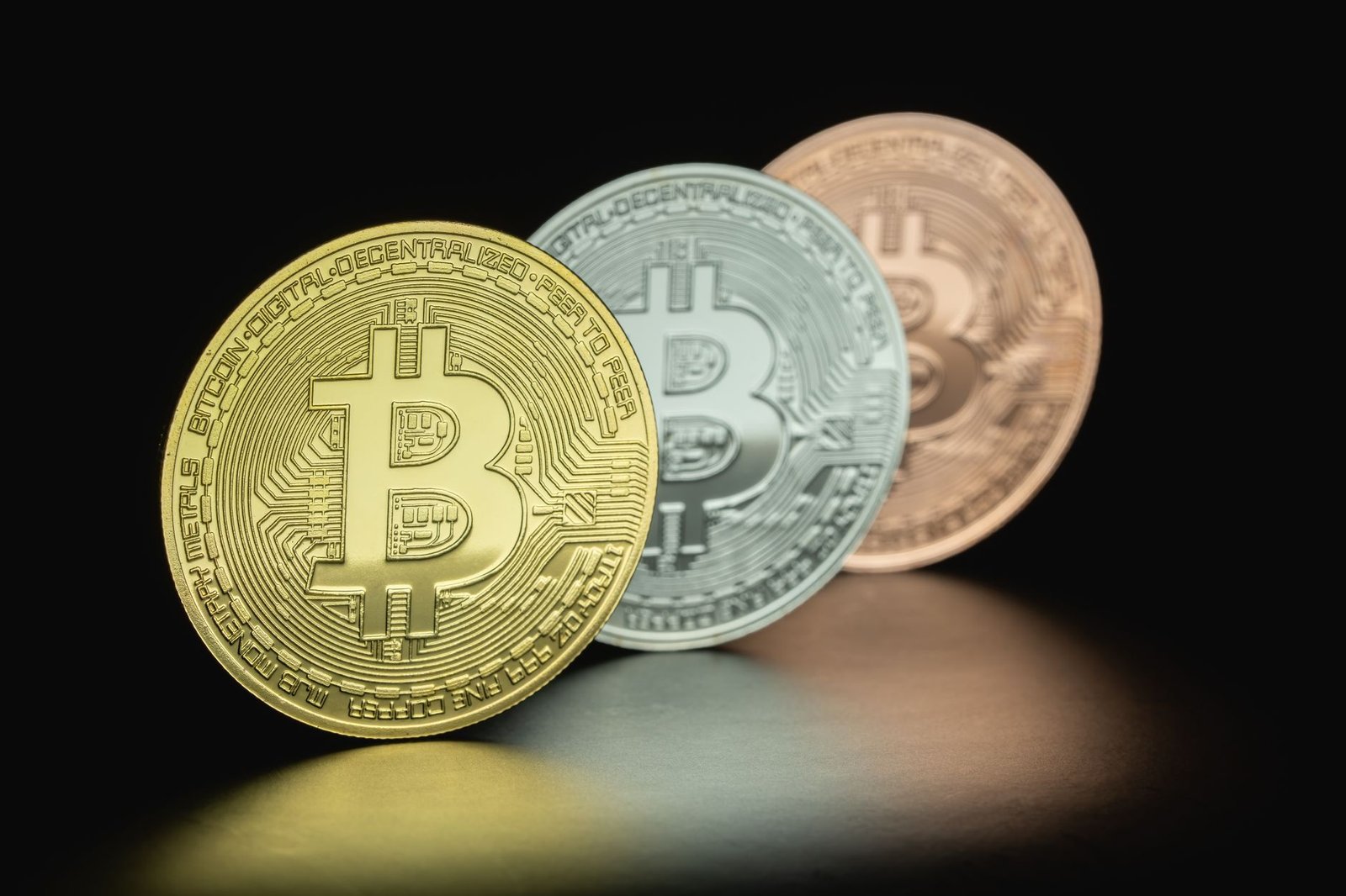 Três fichas metálicas com o símbolo do bitcoin, em dourado, prateado e cobre, sobre fundo escuro