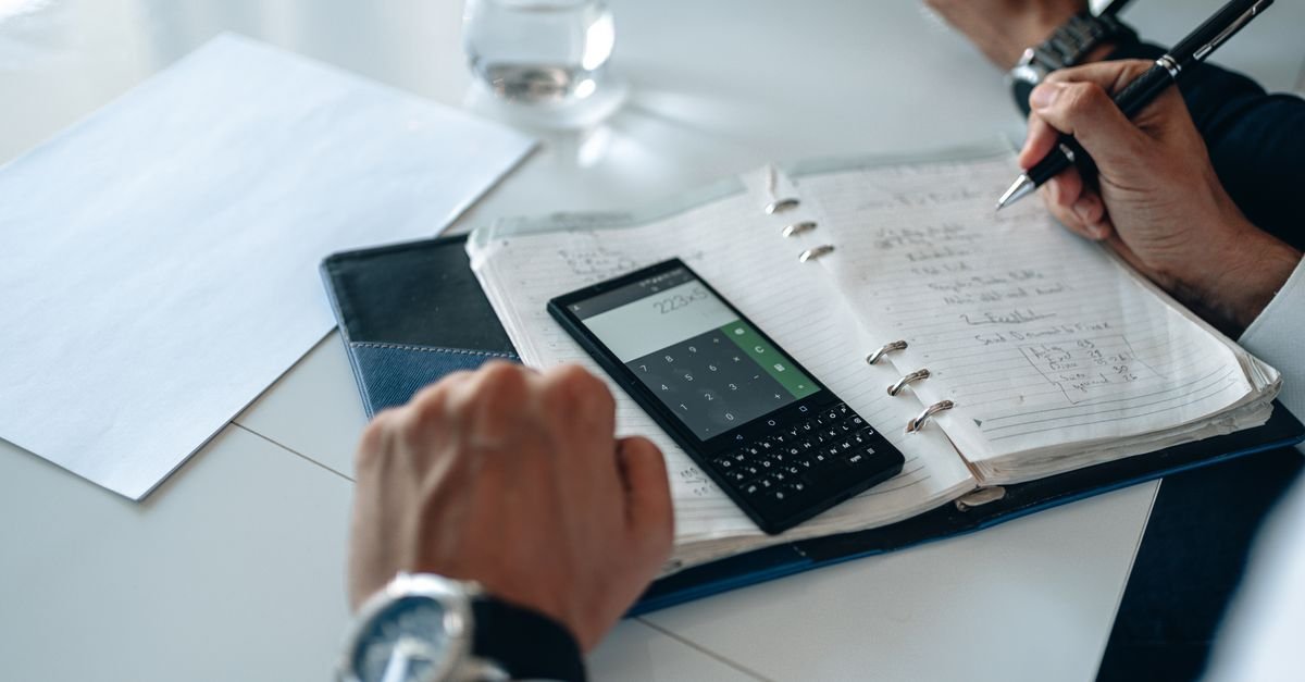 Mãos fazendo contas em agenda de papel com aplicativo de calculadora aberto no celular sobre a mesa