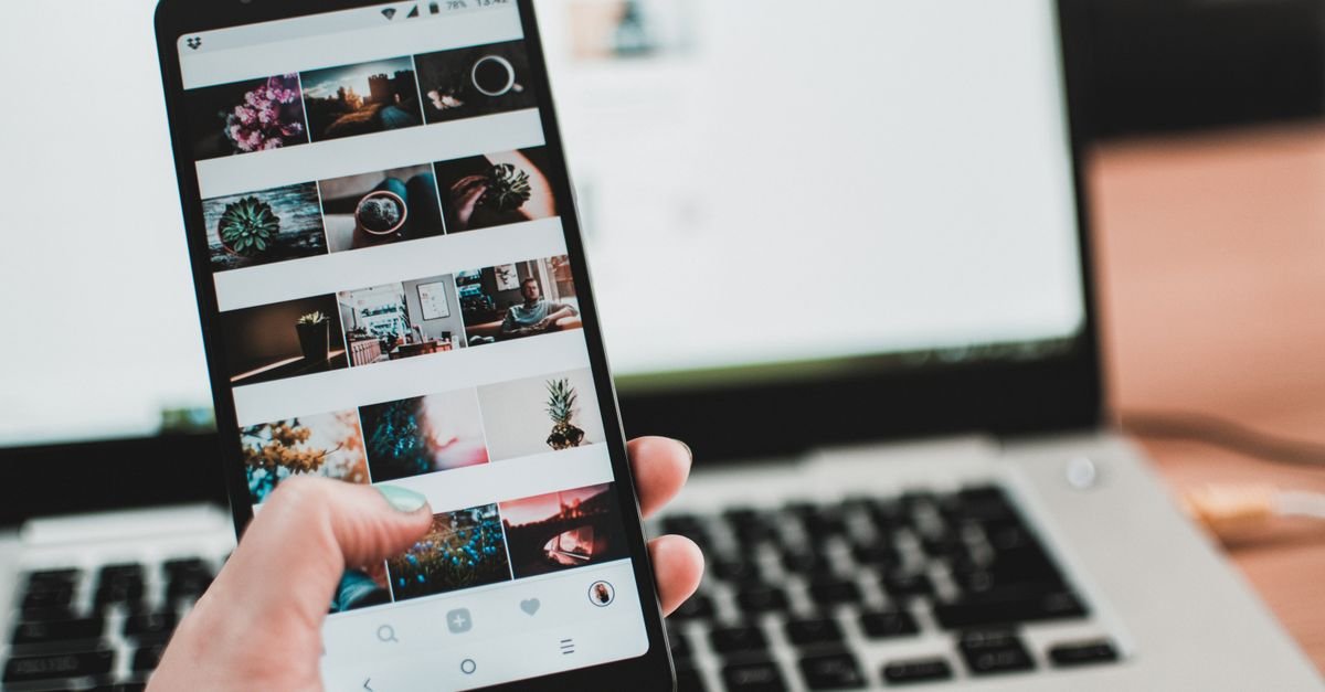 Mão segura um celular com o feed de fotos de uma rede social aberto, diante de um notebook; imagem ilustrativa