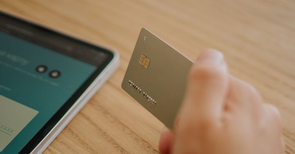 Mão segura cartão bancário dourado ao lado de tablet sobre mesa de madeira