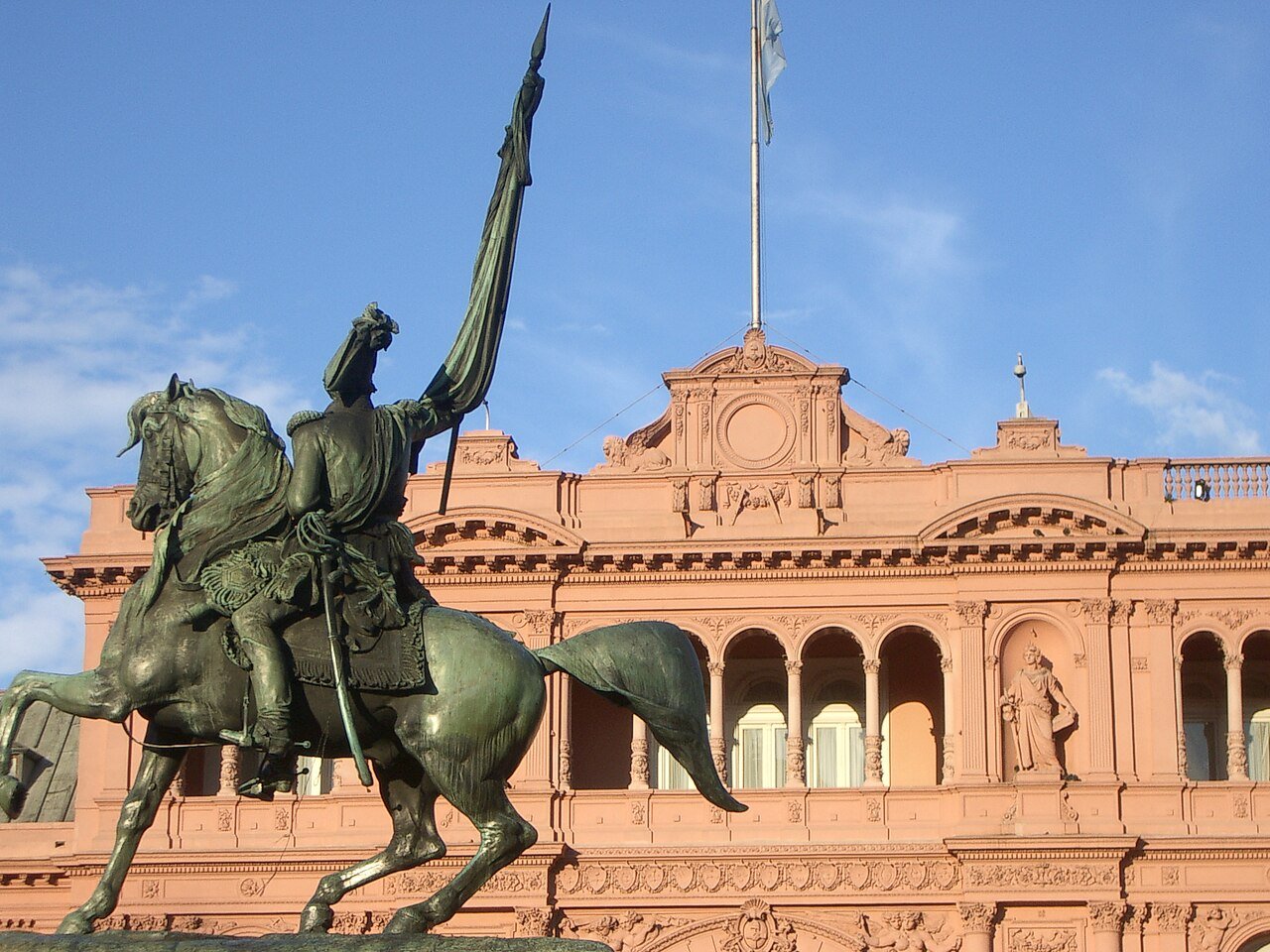 Fachada da Casa Rosada, sede do governo argentino, em Buenos Aires, com a bandeira nacional hasteada e o monumento equestre a Manuel Belgrano em primeiro plano