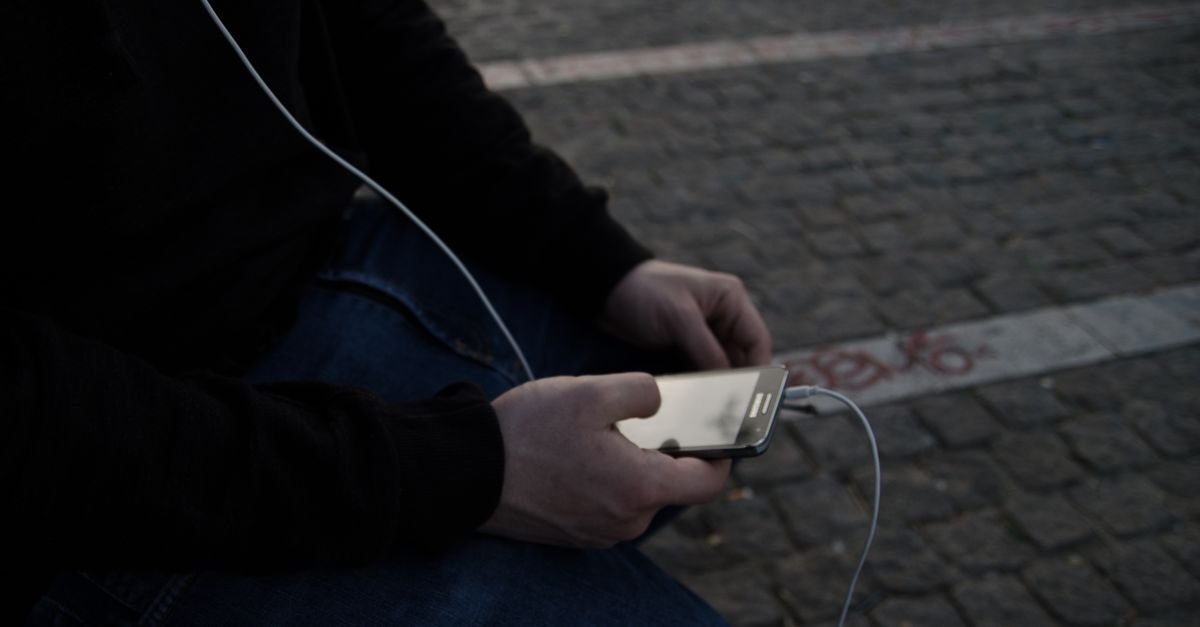 Pessoa sentada na rua segura um celular com as duas mãos