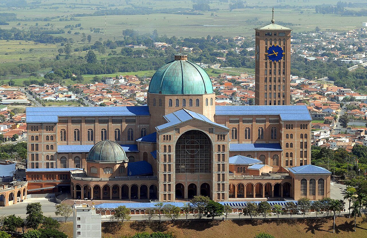 Vista aérea da Basílica Nacional de Nossa Senhora Aparecida, com a cúpula verde e a torre do relógio, cercada pela cidade