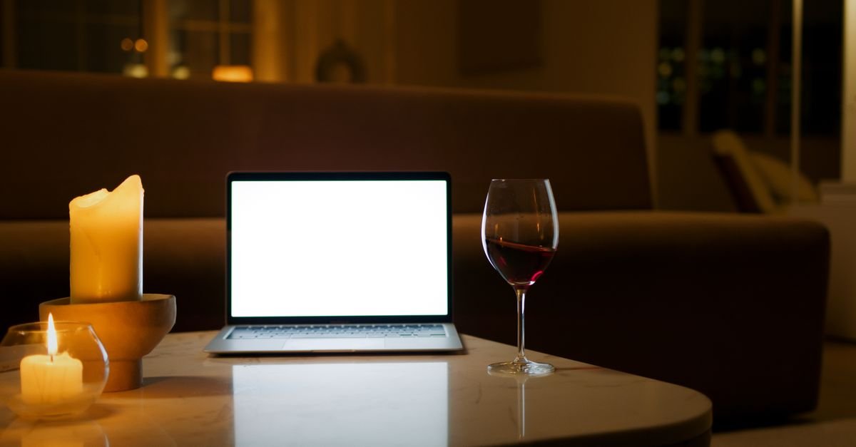 Notebook aberto com tela em branco sobre mesa, ao lado de uma taça de vinho tinto e velas acesas
