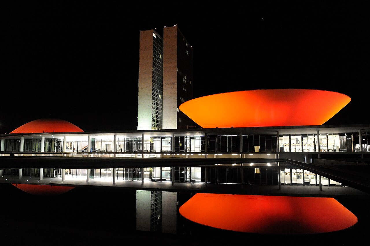 Congresso Nacional à noite, com as cúpulas iluminadas em vermelho refletidas no espelho d'água