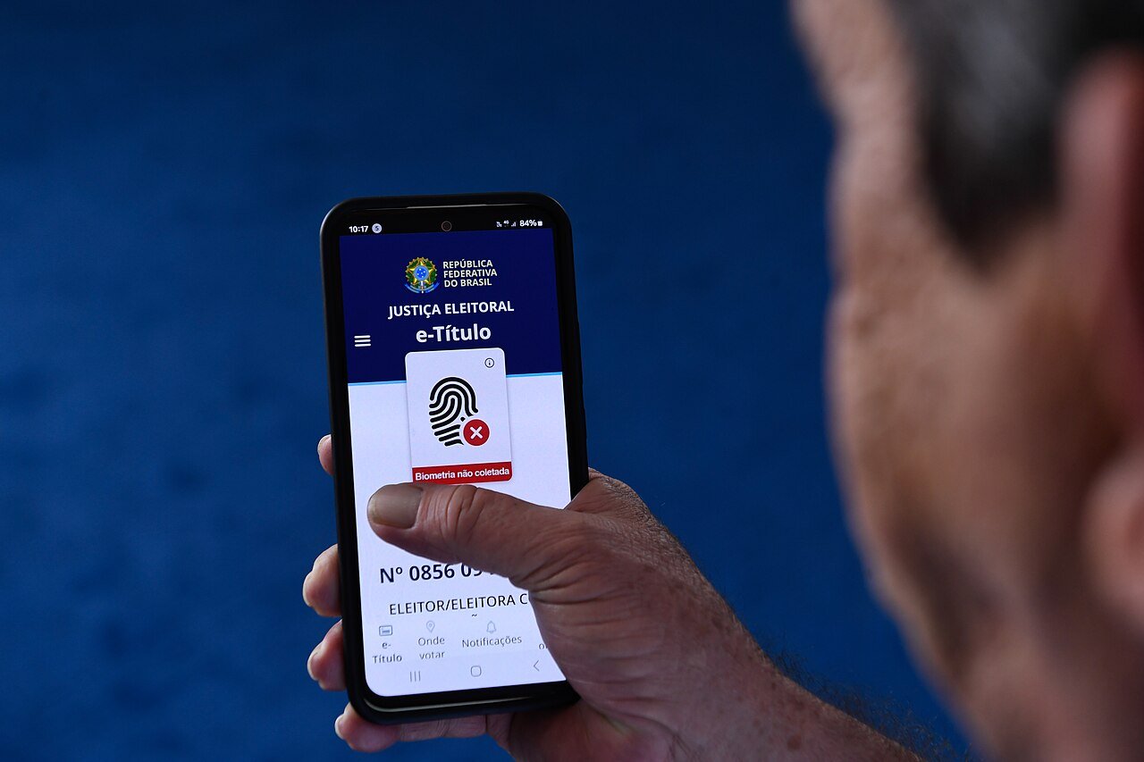 Pessoa segura celular com o aplicativo e-Título aberto, mostrando a aba Onde votar na parte inferior da tela