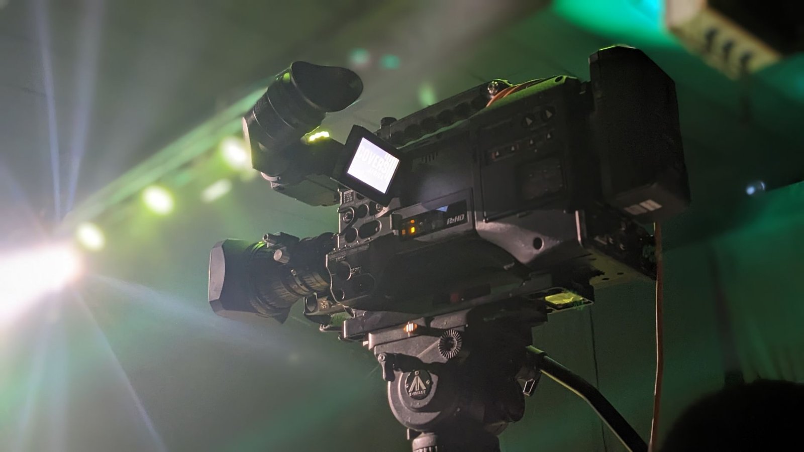 Câmera de televisão profissional apontada para um palco, com refletores acesos ao fundo