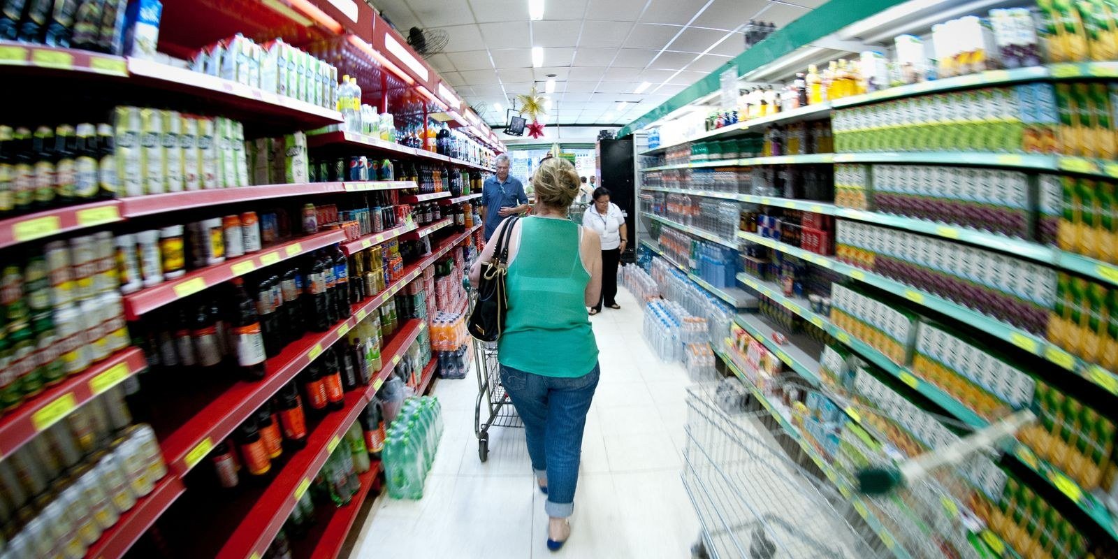 Consumidora empurra carrinho em corredor de supermercado com prateleiras cheias de bebidas e alimentos