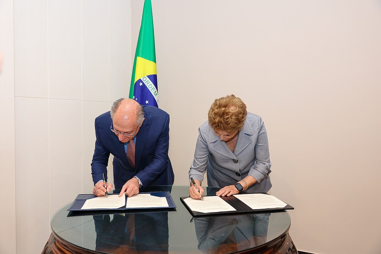 Dilma Rousseff, presidente do Novo Banco de Desenvolvimento, assina documento ao lado do vice-presidente da República em mesa com a bandeira do Brasil ao fundo, em junho de 2024