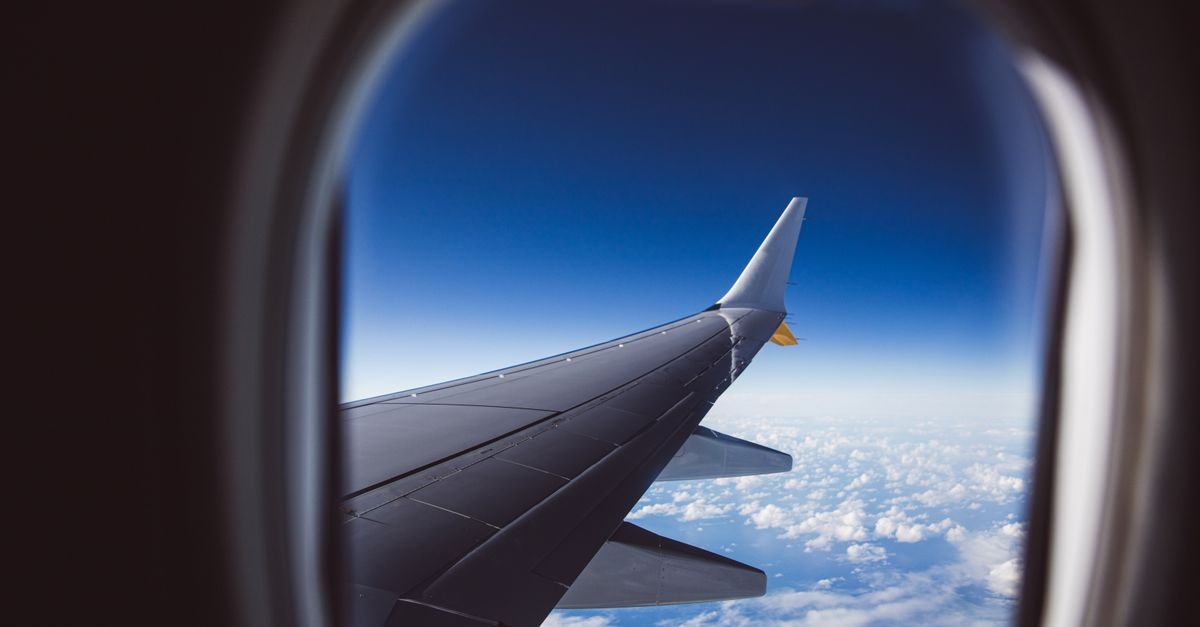 Asa de um aviao vista pela janela da cabine, com ceu azul e nuvens abaixo