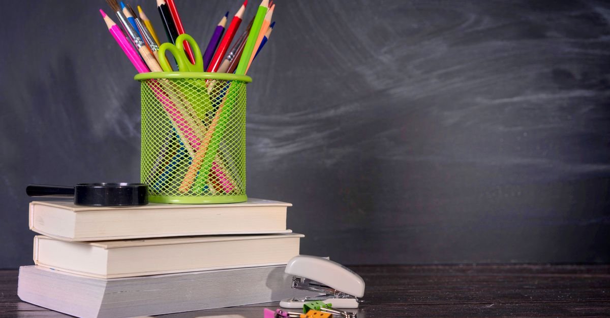 Material escolar com lápis, livros e grampeador sobre mesa diante de um quadro-negro