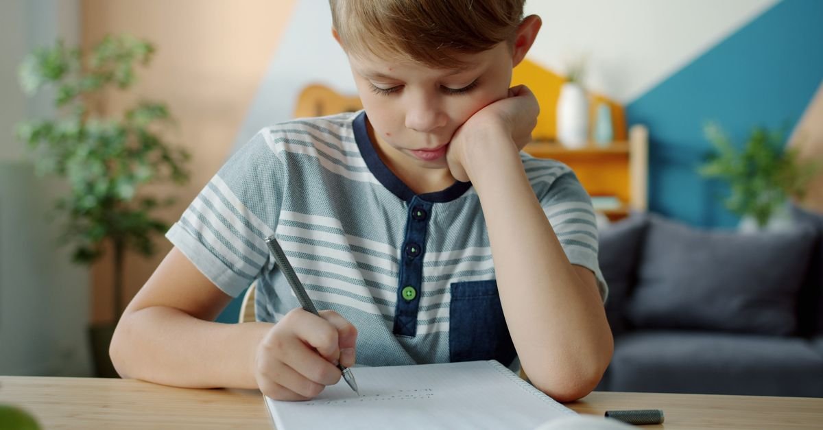 Menino faz atividade escolar em caderno sobre a mesa, em casa