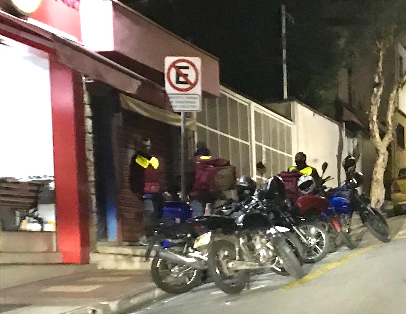Motocicletas de entregadores estacionadas em fila na calçada em frente a um estabelecimento comercial à noite