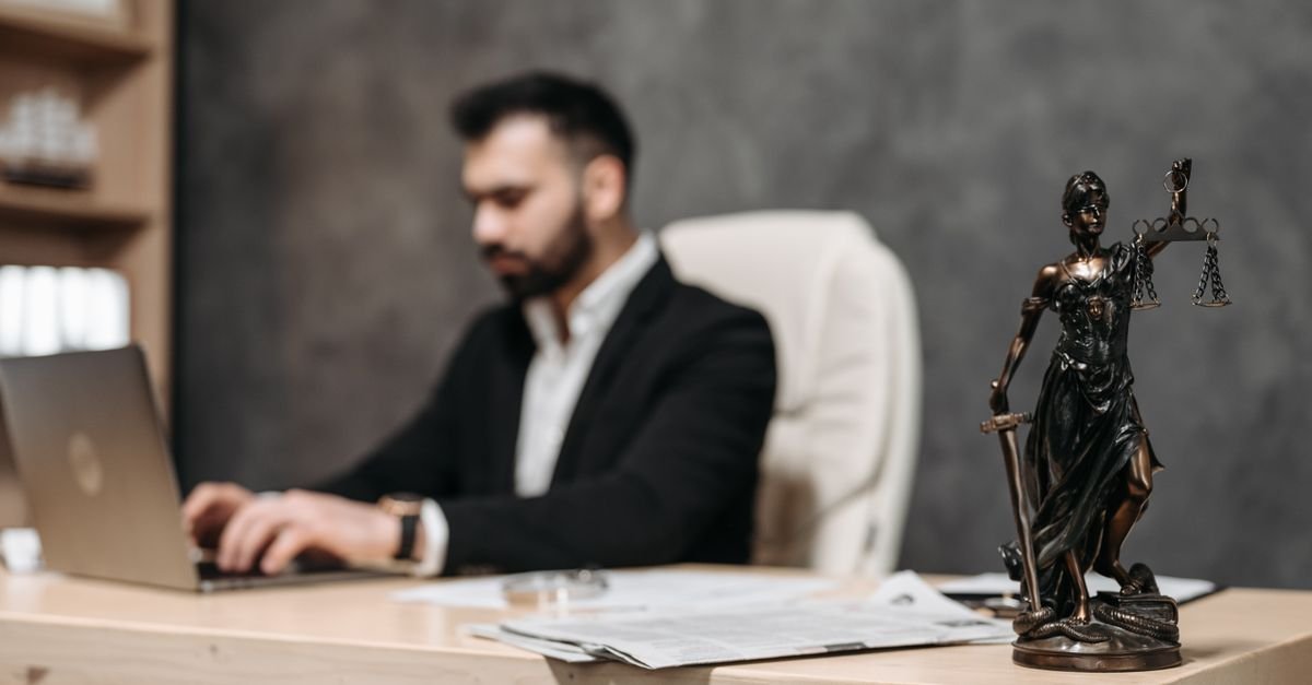 Estátua da Justiça com balança sobre mesa de escritório, com homem trabalhando em notebook ao fundo