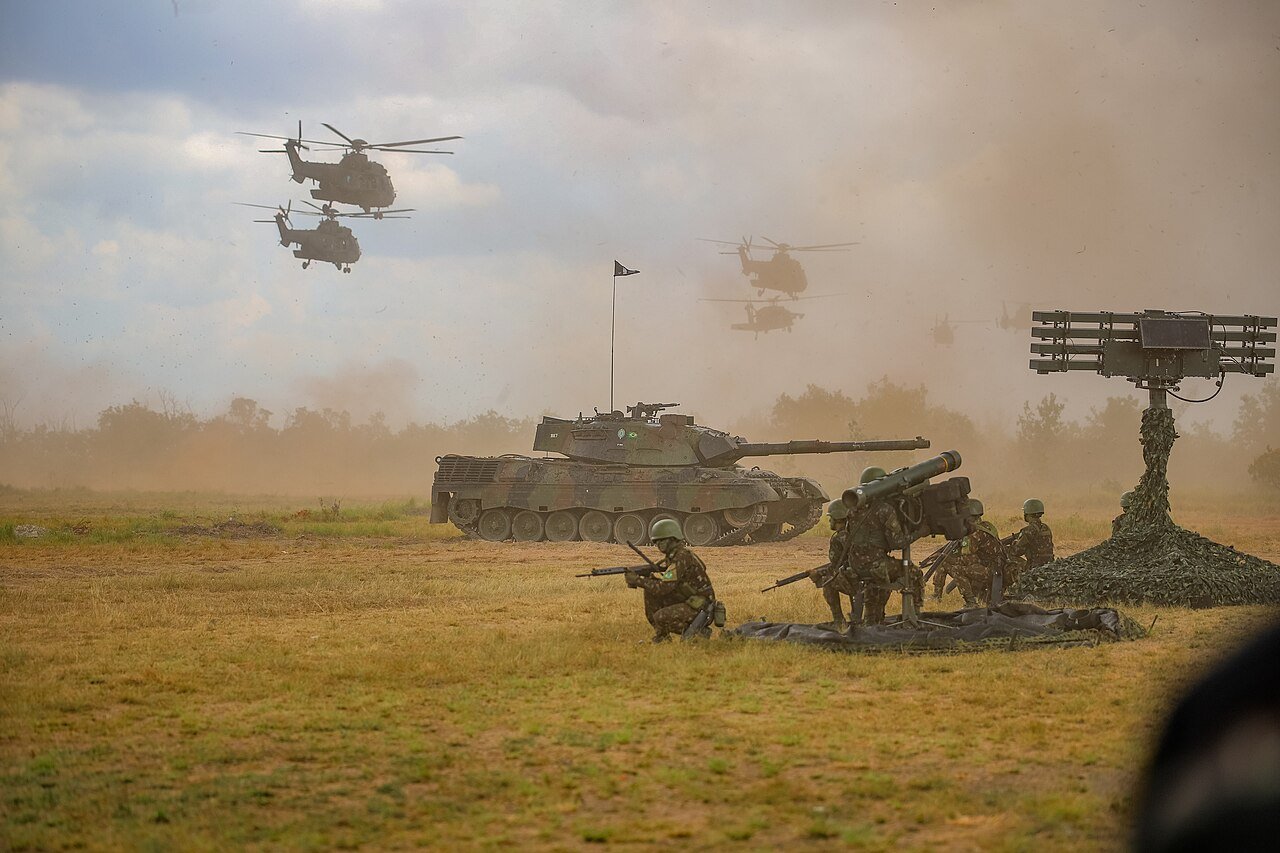 Militares do Exército Brasileiro operam lançador antiaéreo e radar em campo aberto, com blindado e helicópteros ao fundo durante exercício