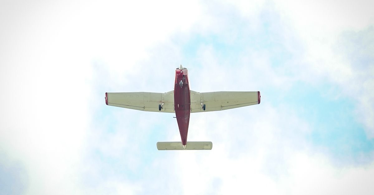 Avião monomotor de pequeno porte visto de baixo, voando em céu com nuvens