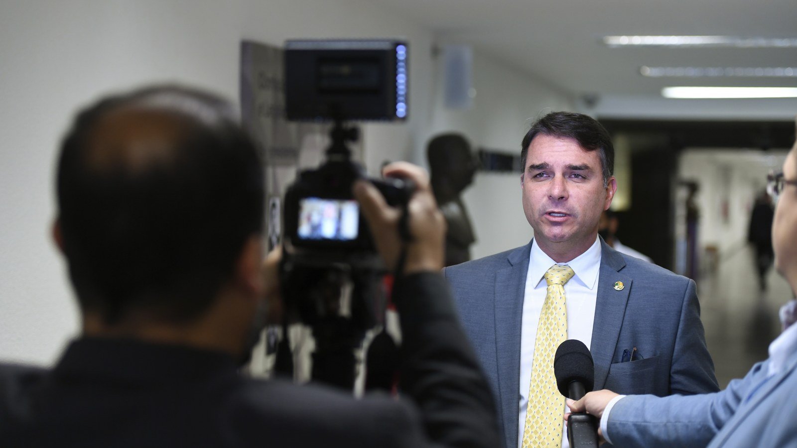 Senador Flávio Bolsonaro de terno cinza e gravata amarela concede entrevista em corredor do Senado, diante de câmera e microfone