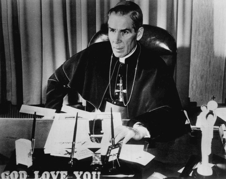 O arcebispo Fulton Sheen sentado à sua mesa de trabalho em registro de 1956