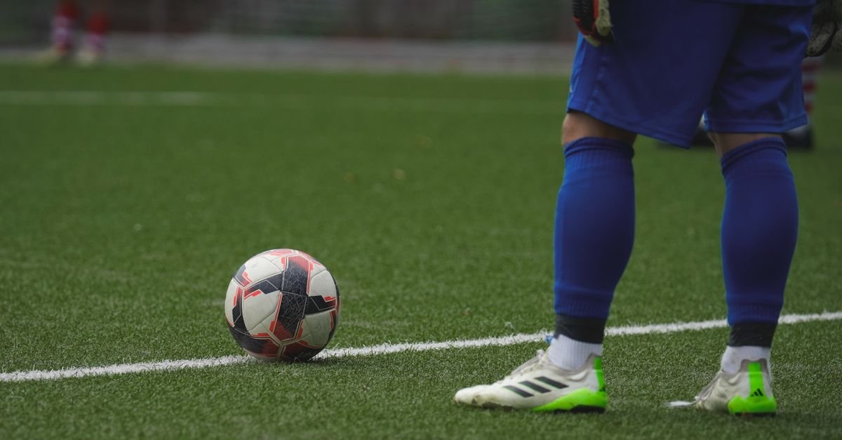 Bola de futebol parada no gramado ao lado das pernas de um jogador