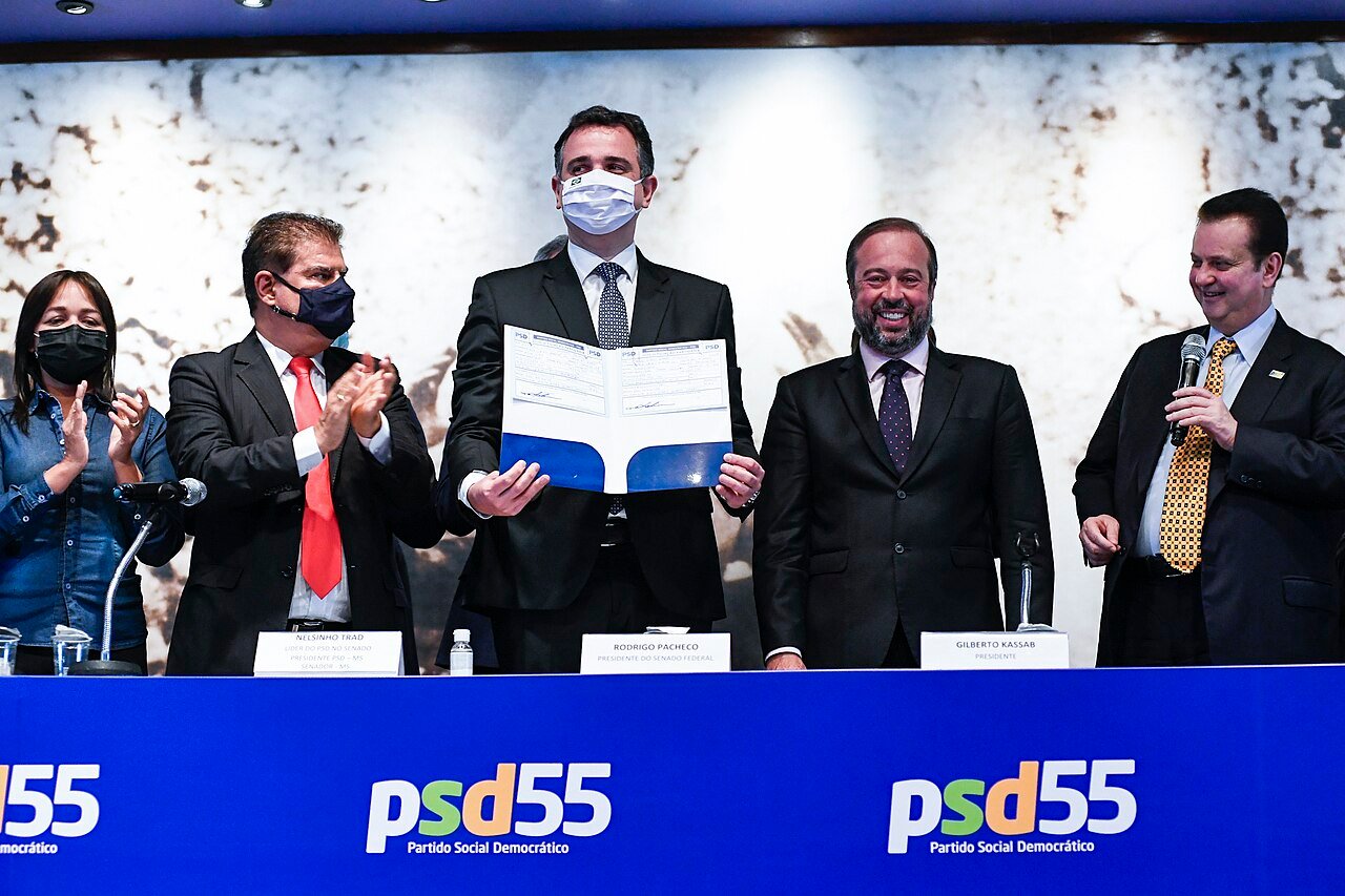Gilberto Kassab fala ao microfone em evento do PSD, ao lado de outros dirigentes, diante de painel com a marca do partido
