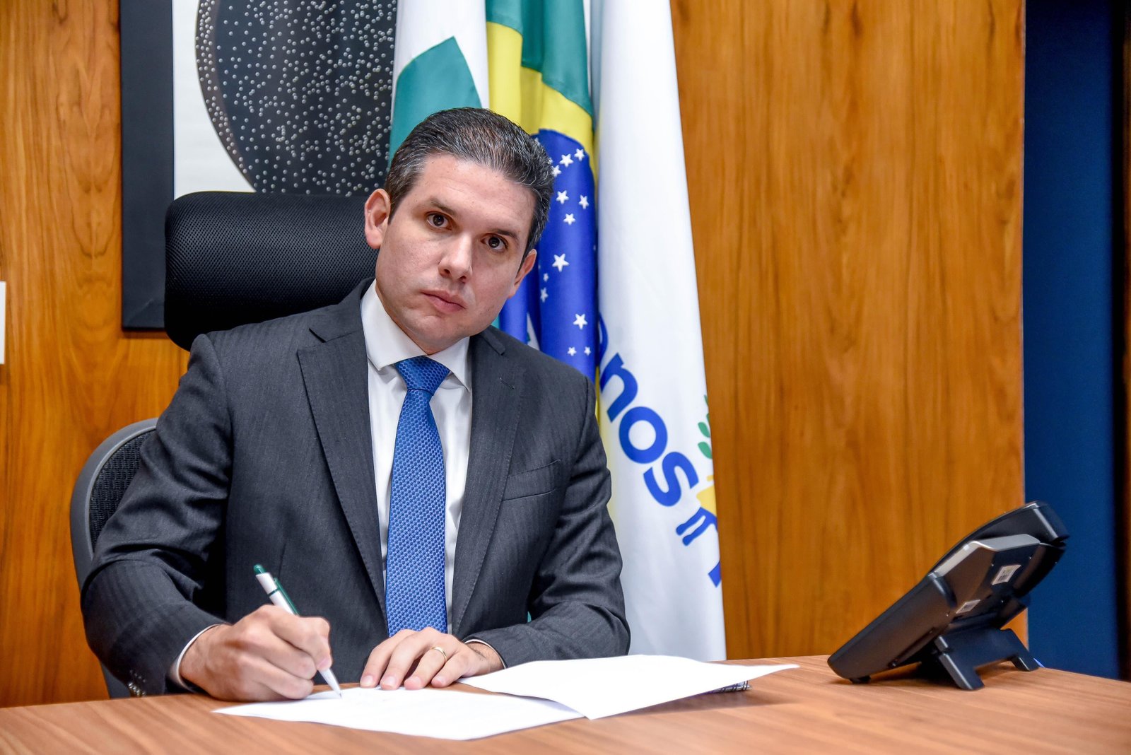 Hugo Motta sentado à mesa de gabinete assinando documento, com bandeiras do Brasil e da Paraíba ao fundo