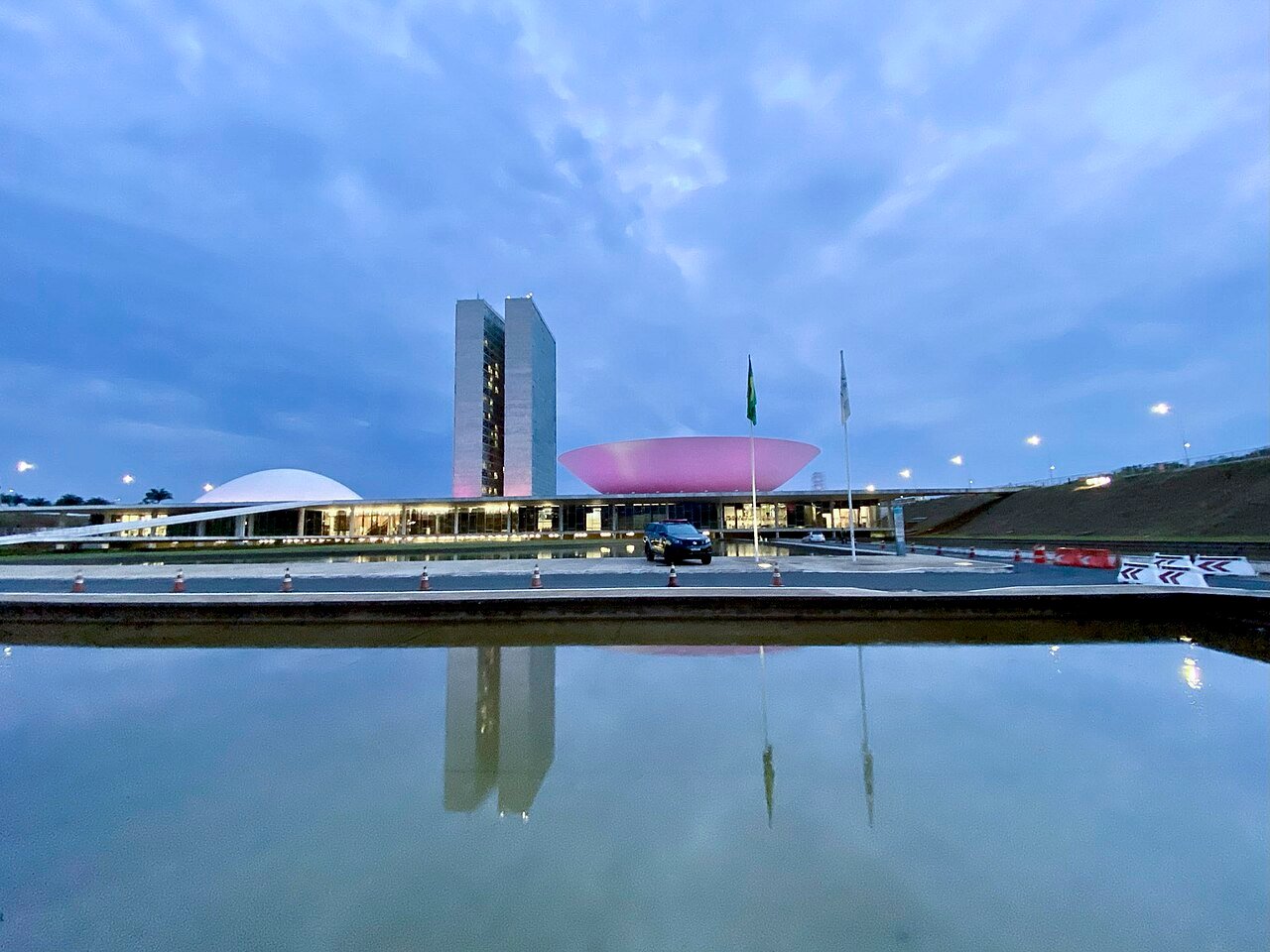 Fachada do Congresso Nacional no fim da tarde, com a cúpula do Senado iluminada e o espelho dágua em primeiro plano