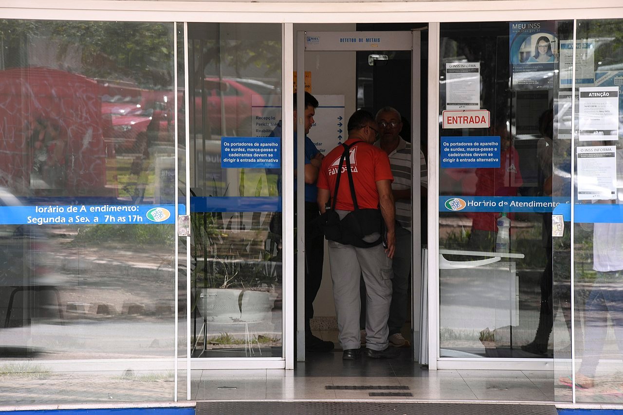 Entrada de vidro de uma agencia do INSS com cartazes de atendimento e pessoas na porta