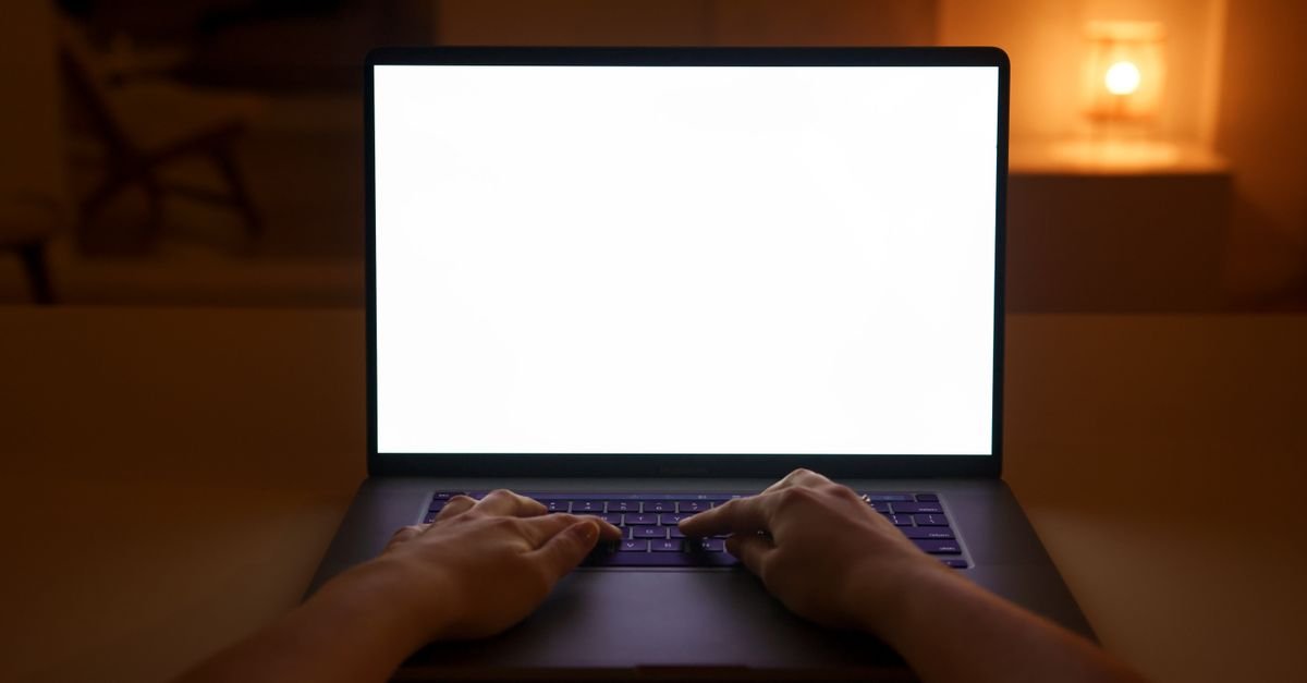 Mãos digitando em teclado de notebook com a tela acesa em branco, em ambiente escuro iluminado por abajur