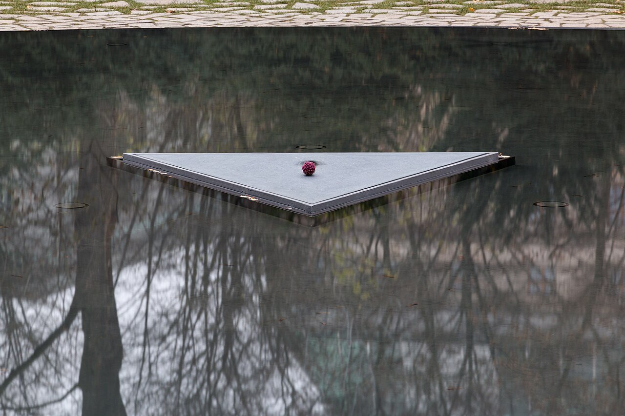 Estela triangular de pedra no centro do espelho d'água do memorial de Berlim às vítimas ciganas do nazismo, com uma flor sobre a superfície