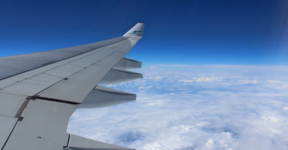 Asa de avião em voo vista de dentro da cabine, acima de uma camada de nuvens
