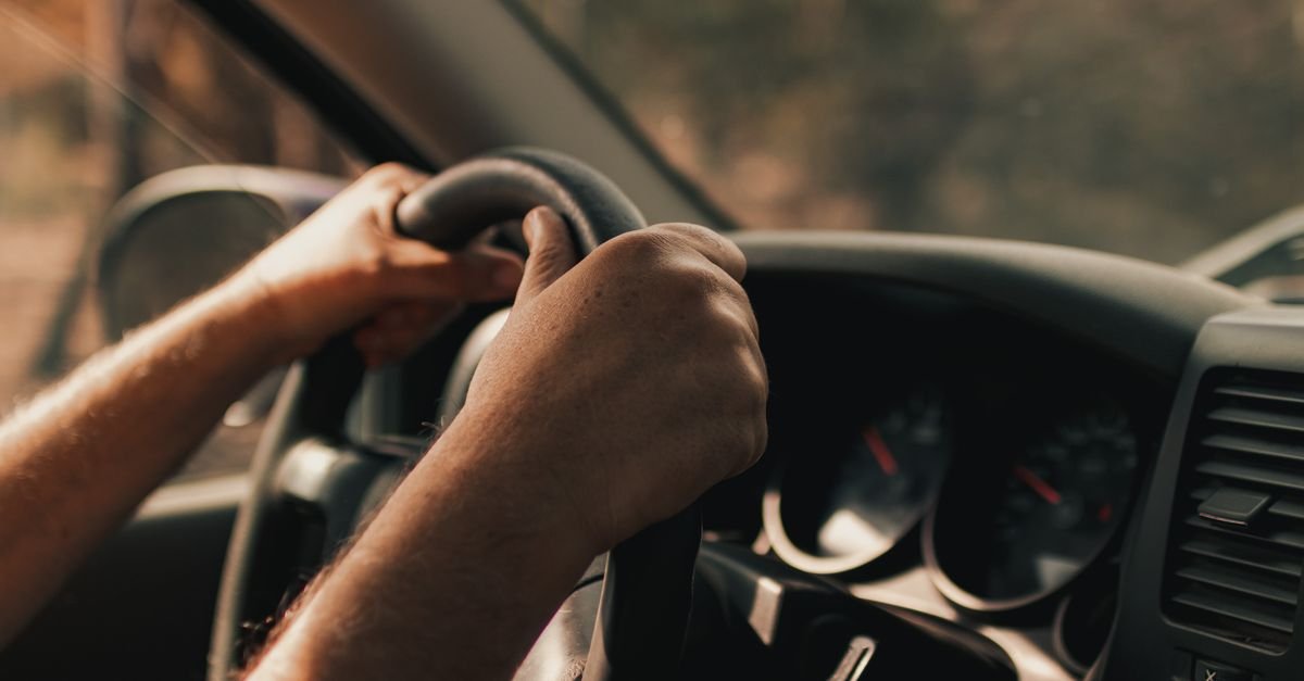Mãos de motorista no volante de um carro em movimento, com painel do veículo ao fundo