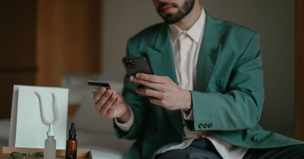 Homem sentado segura cartão de crédito e celular para fazer compra pela internet