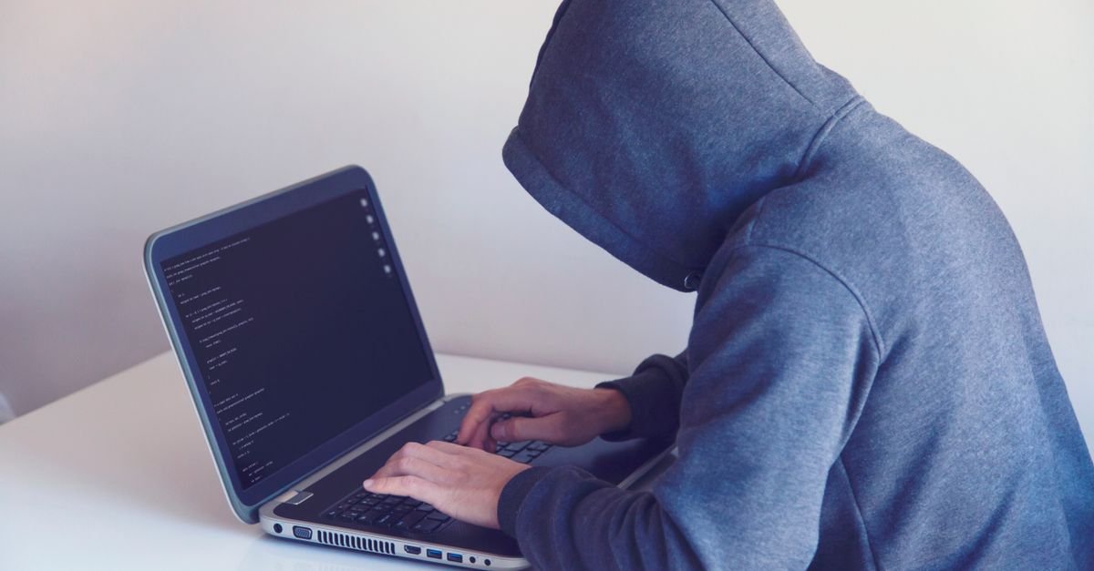 Pessoa de capuz cinza digitando em notebook com linhas de código na tela