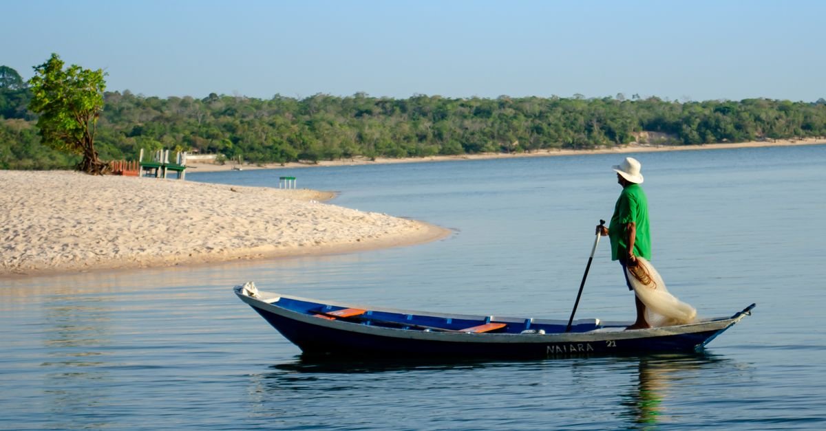 Pescador em pé numa canoa de madeira recolhe a rede em rio de águas calmas
