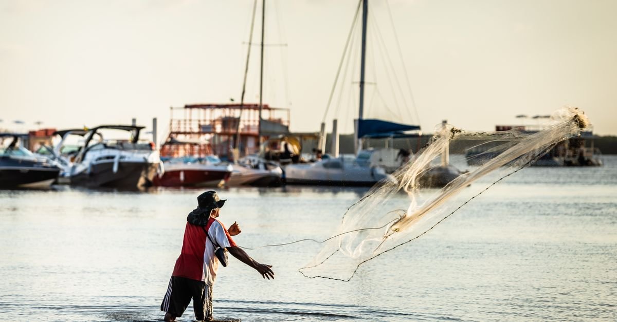 Pescador lança tarrafa em águas calmas de rio no Brasil, com embarcações ao fundo
