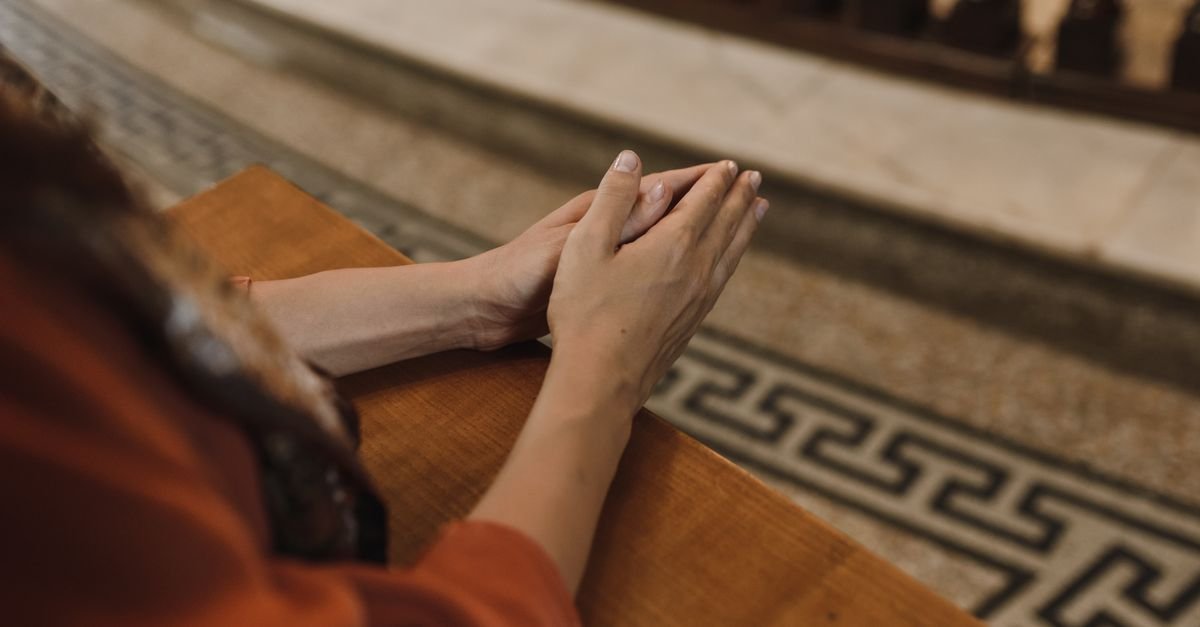 Mãos unidas em oração apoiadas em banco de madeira dentro de uma igreja