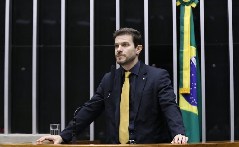 Deputado Felipe Becari na tribuna do plenário da Câmara dos Deputados ao lado da bandeira do Brasil