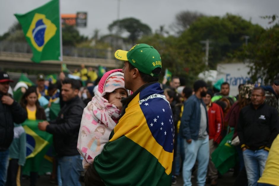 Homem com boné verde e amarelo carrega uma criança embrulhada em bandeira do Brasil em ato ao ar livre