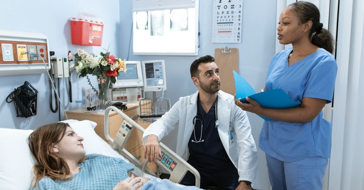 Médico e enfermeira conversam ao lado do leito de uma paciente em quarto de hospital