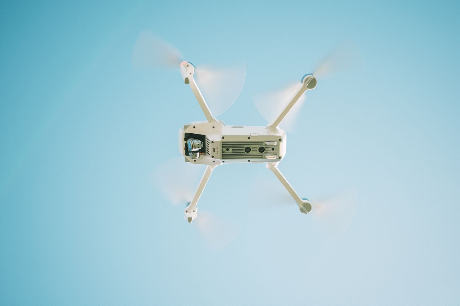 Drone branco em voo visto de baixo contra o céu azul, com as hélices em movimento