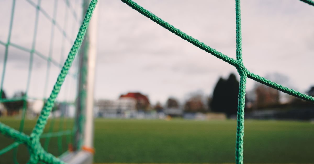 Rede de gol de futebol em primeiro plano, com o gramado do campo ao fundo