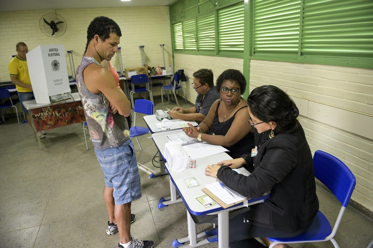 Eleitor apresenta documento na mesa receptora de votos, com cabine de votação da Justiça Eleitoral ao fundo