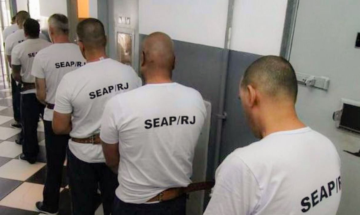 Fila de custodiados de costas com camisetas da Secretaria de Administracao Penitenciaria do Rio de Janeiro