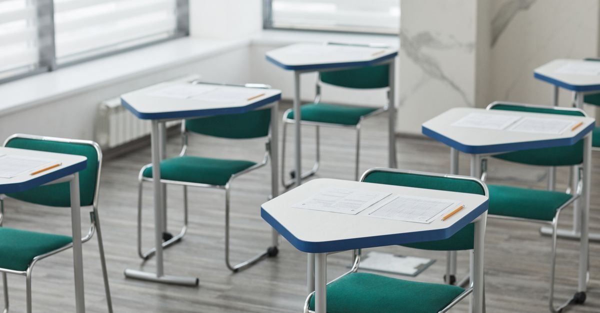 Sala de aula vazia com carteiras individuais enfileiradas e folhas de exercício sobre as mesas