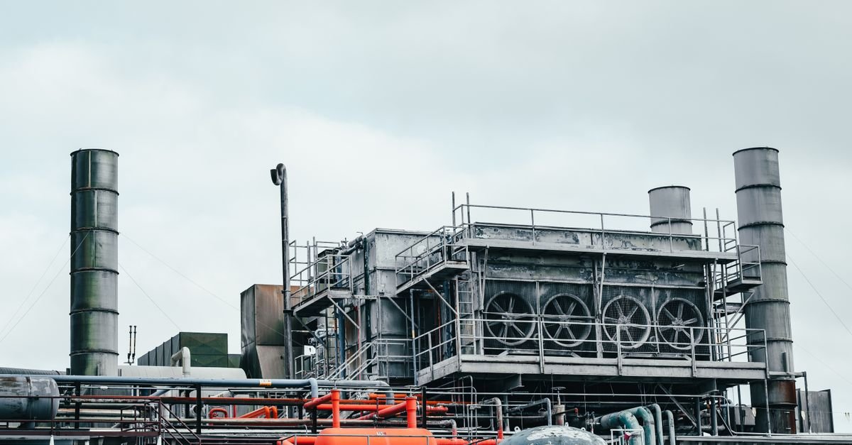 Planta industrial com tubulações, chaminés e equipamentos metálicos sob céu nublado