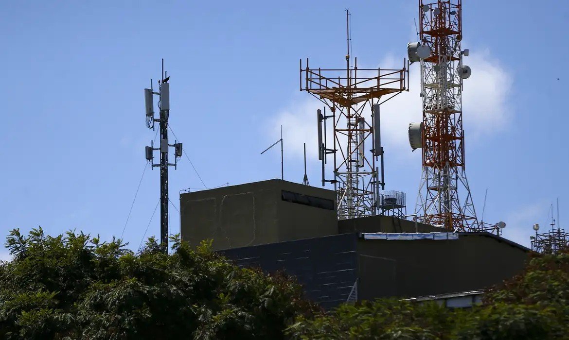 Torres de antenas de telecomunicações instaladas sobre a laje de um prédio, com céu azul ao fundo